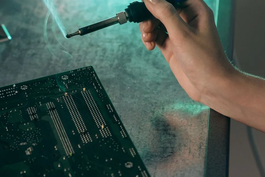 Soldering iron working on a handheld console motherboard circuit board with wires and flux